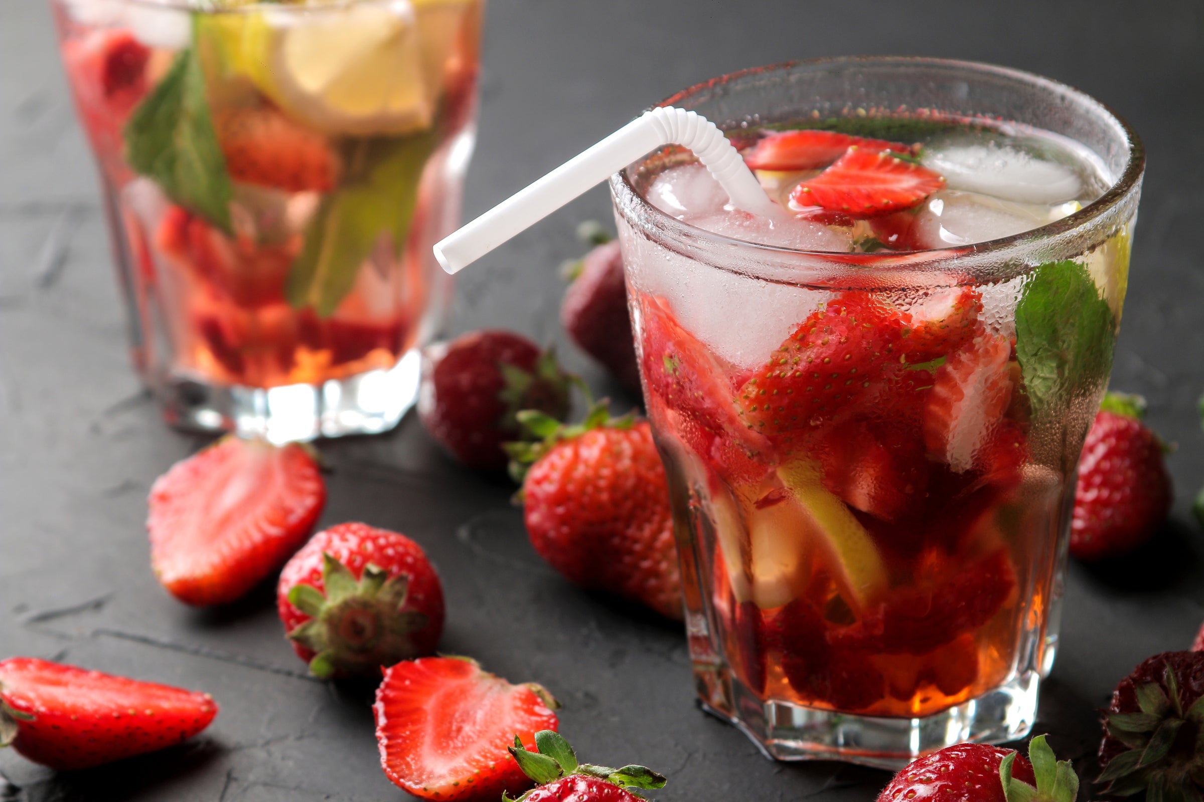 Glas mit Eistee und vielen Erdbeerstückchen sowie Zitronenscheiben. Mit Eiswürfel und Strohhalm (altes Foto noch mit Plastikstrohhalm). Im Vordergrund liegen weitere Erdbeeren und im Hintergrund ein zweites Glas unscharf.