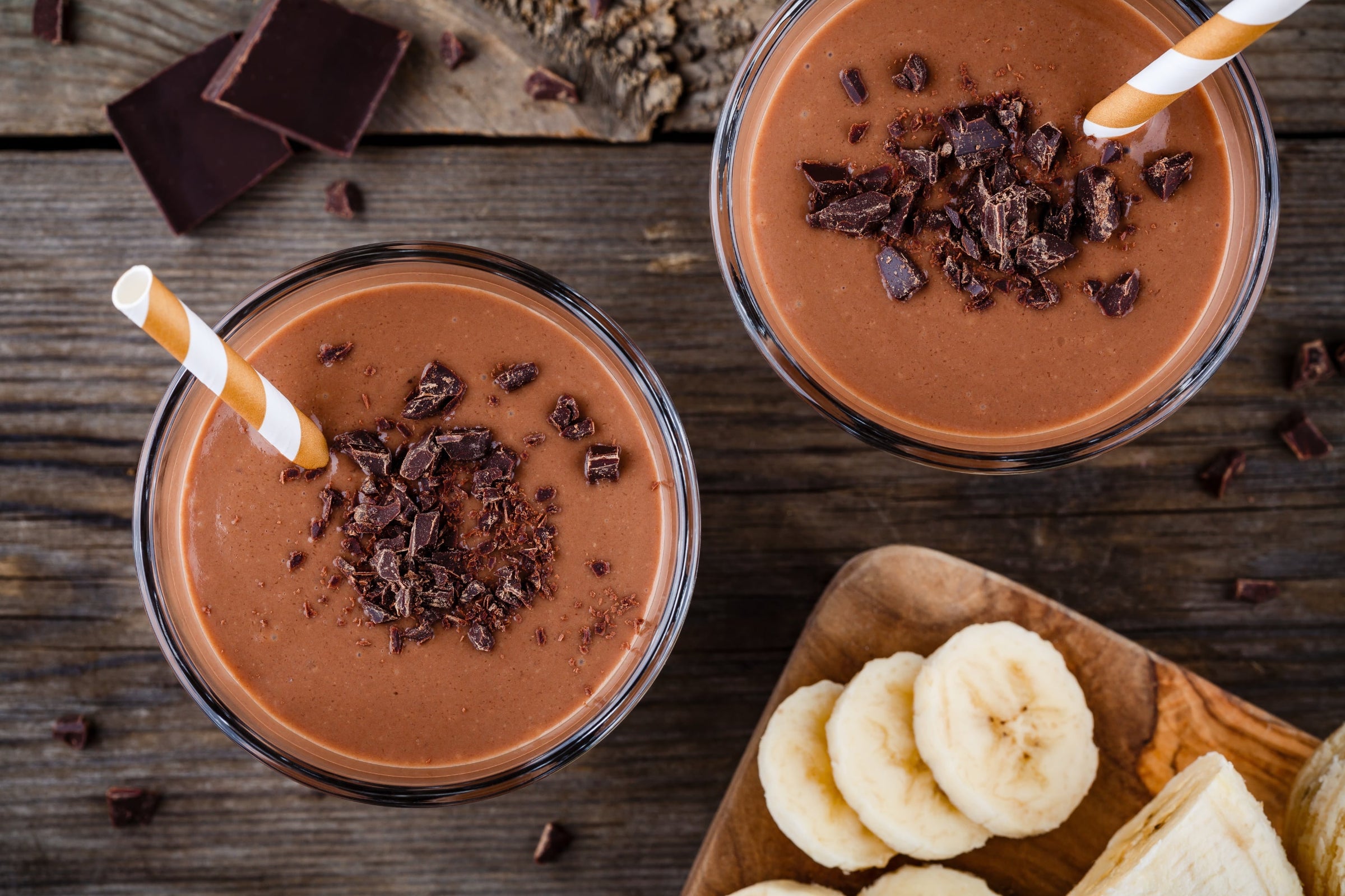2 Rooibos Bananen Schoko Shakes im Glas von oben fotografiert mit Papier-Röhrchen und Schoko Stückchen auf einem Tisch. Schokostücke und geschnitten Bananen sind ebenso zu sehen.